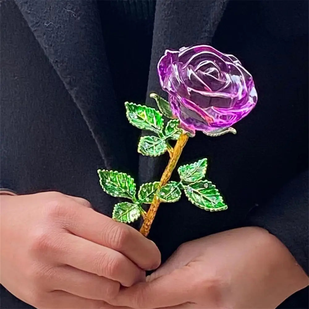 A translucent purple crystal glass rose with green enamel leaves and a gold-toned stem held in hands against dark fabric for a Valentine’s Day gift.