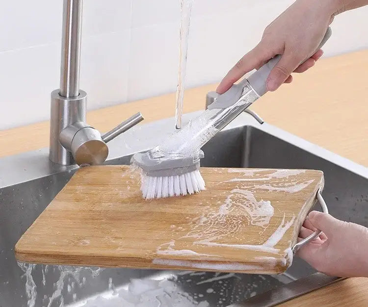 Brosse à vaisselle pour distribution de savon en plastique grise et transparente avec brins blancs et long manche, conçue pour récurer une planche à découper en bois sous l’eau courante.