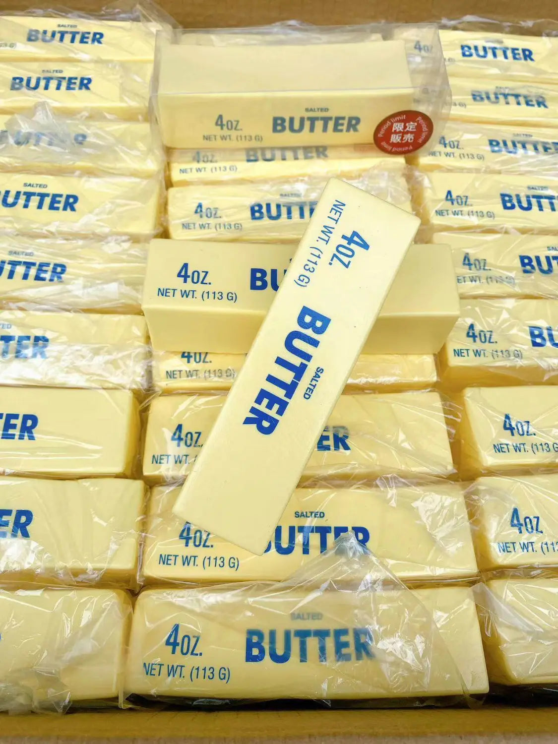Un bloque rectangular de mantequilla salada, de color amarillo pálido, envuelto en plástico transparente con texto impreso azul mostrado desde el frente como un juguete antiestrés blando que asciende lentamente.