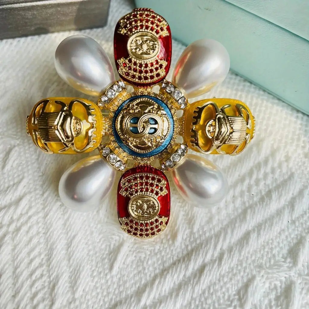 Broche de perles designer présentant une forme florale vibrante, des motifs de coléoptères à ton doré et des pétales d’émail rouge et jaune, avec des détails minutieux.