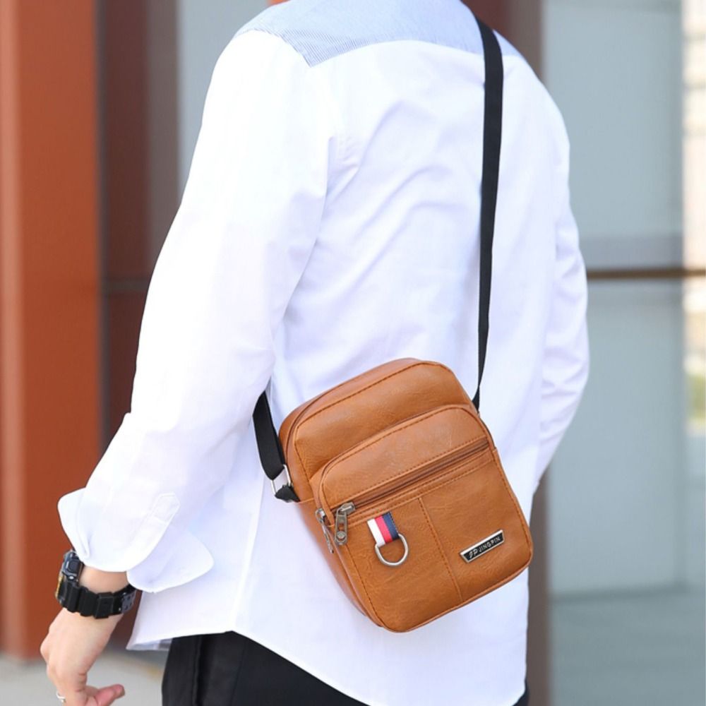 Hellbraune Ledertasche im Sling-Bag-Stil mit schwarzem Gurt und kleinem Flaggenemblem am vorderen Reißverschluss-Zipper, entworfen als legere Umhängetasche im Schulter-Design, Crossbody-Tasche für Herren