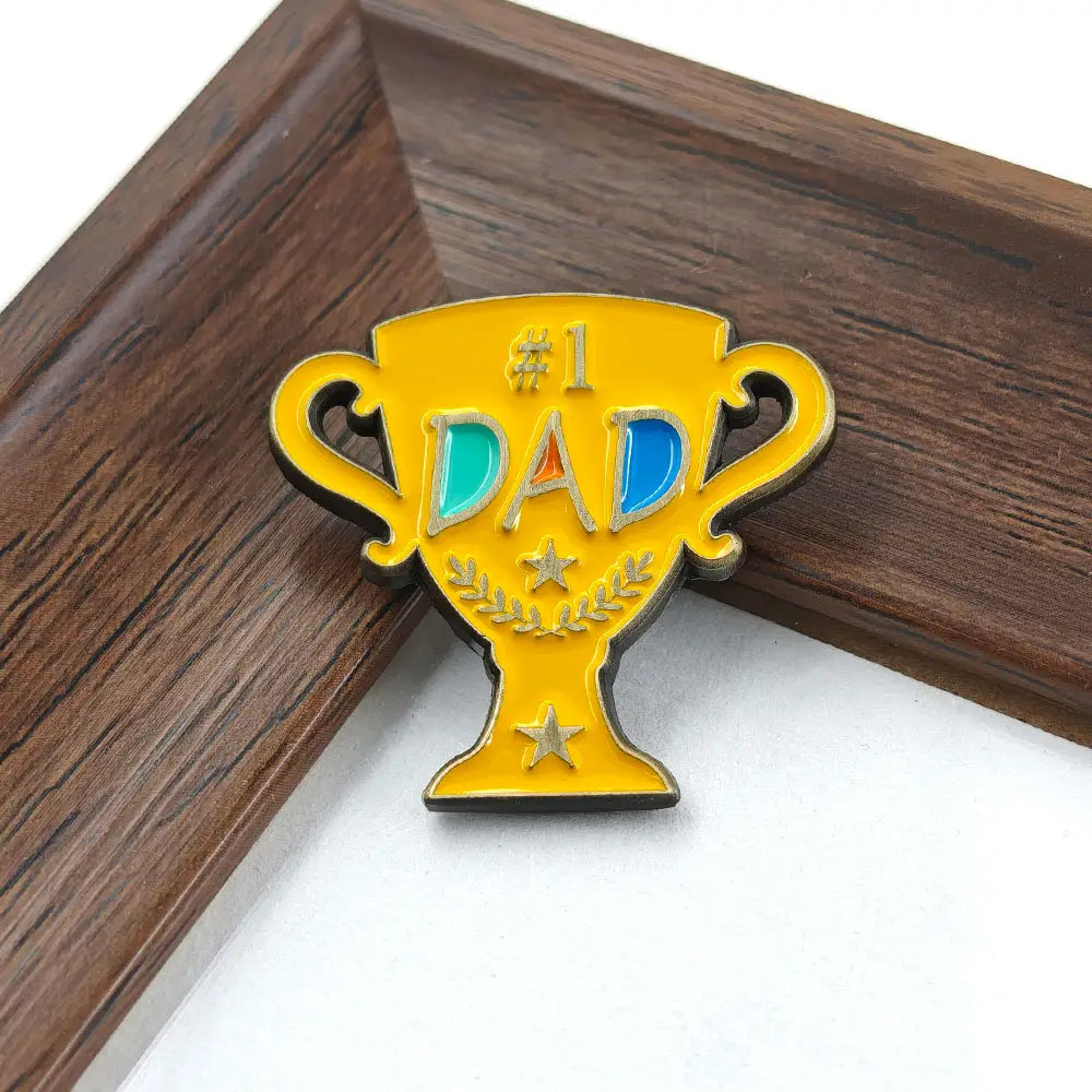 Broche en émail jaune en forme de trophée avec « #1 Dad » gravé en lettres bleues et argentées, rehaussé d’étoiles dorées et de détails dorés de feuilles de laurier sur une broche dorée en émail de trophée « #1 Dad ».