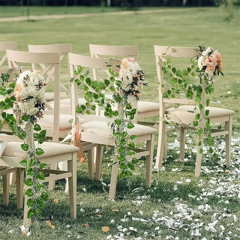 Beigefarbene Holzstühle, dekoriert mit einer grünen künstlichen Pflanzenrankengewächs- Efeublatt-Girlande und floralen Arrangements, von vorn betrachtet.