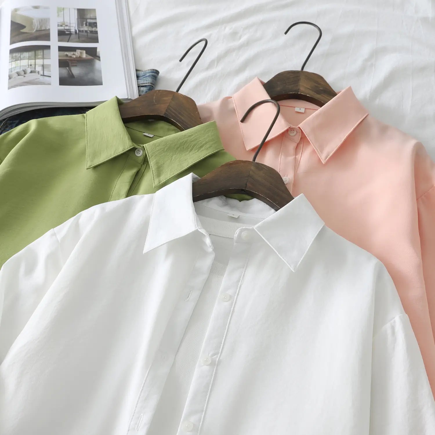 Drei Damen-Casualblusen aus Baumwollmischgewebe mit langen Ärmeln in Weiß, Hellgrün und hellem Rosa, die an Holzkleiderbügeln hängen.