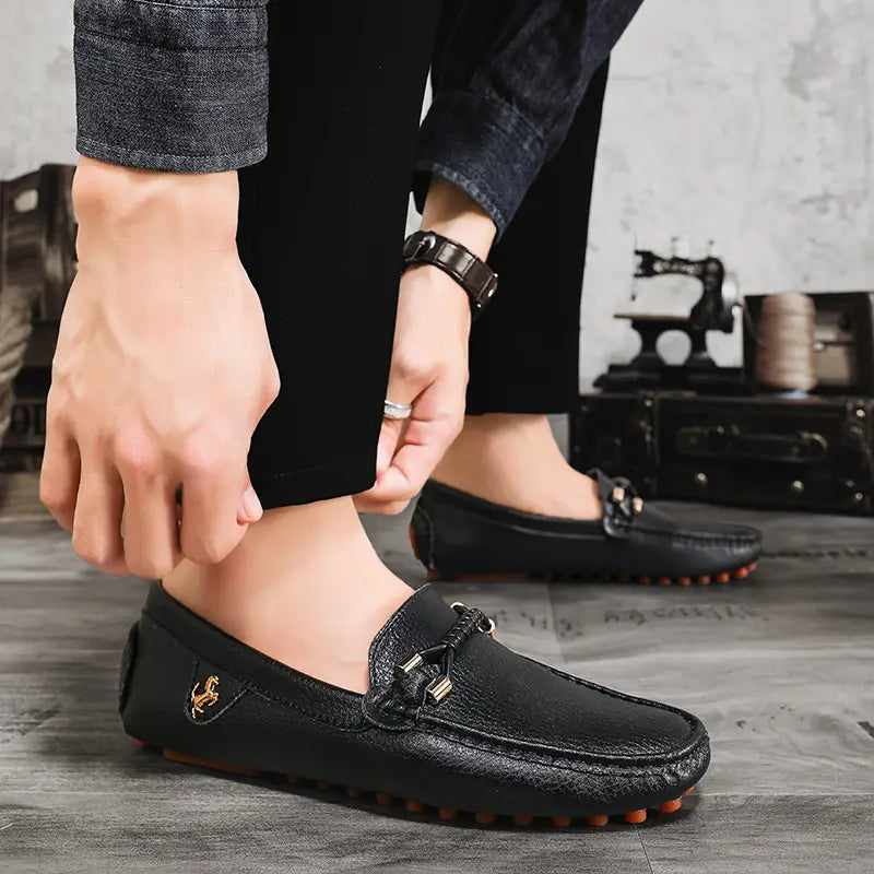 Les chaussures à enfiler habillées en cuir se portent sans effort avec des chaussures décontractées en cuir noir, avec une surface texturée, un emblème équestre doré sur le côté et un détail de boucle métallique.