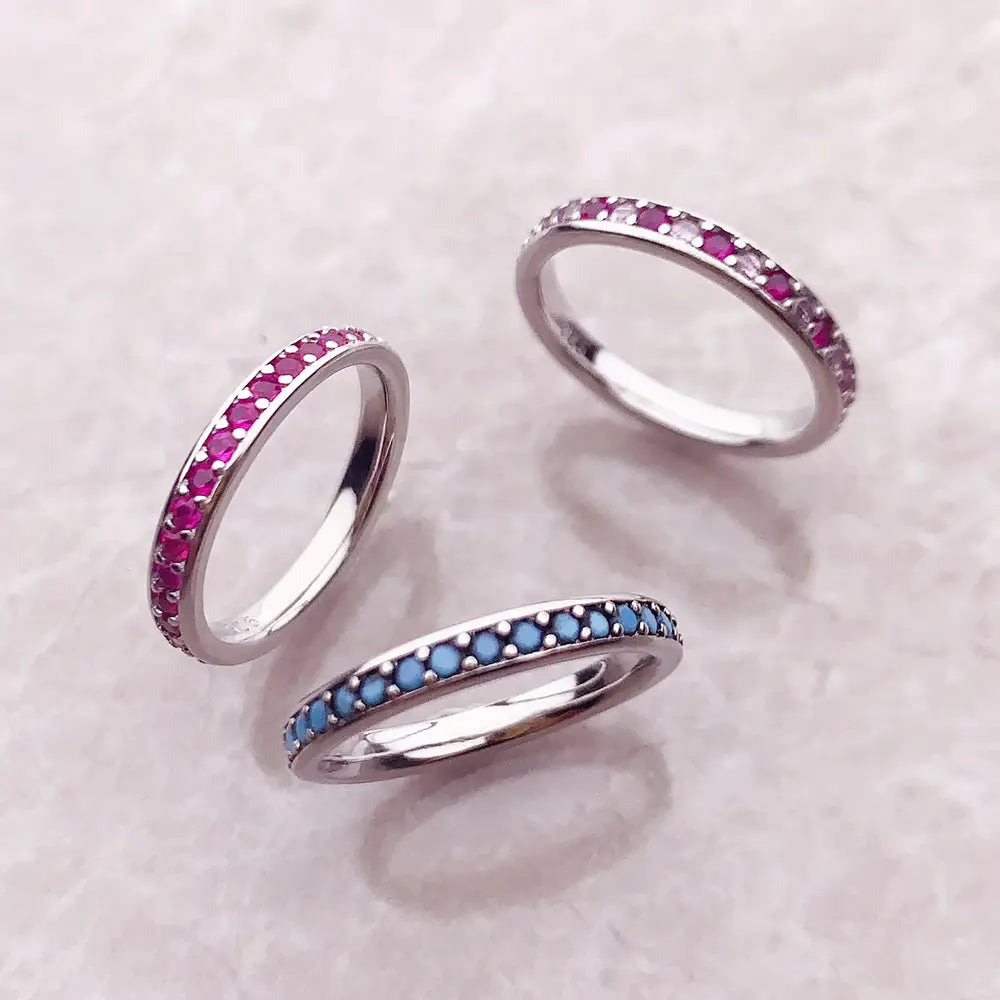 Trois délicates bagues empilées en argent sterling, avec des rangées continues de rubis roses vibrants, de pierres bleues, et des cristaux alternant entre rose et transparent.