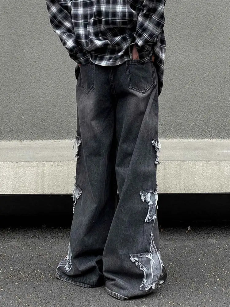 Jeans denim Y2k baggy délavé et déchiré gris foncé fabriqué en denim, présentant des patchs effilochés et déchirés ainsi qu’une déchirure en forme d’étoile sur la partie inférieure de la jambe.