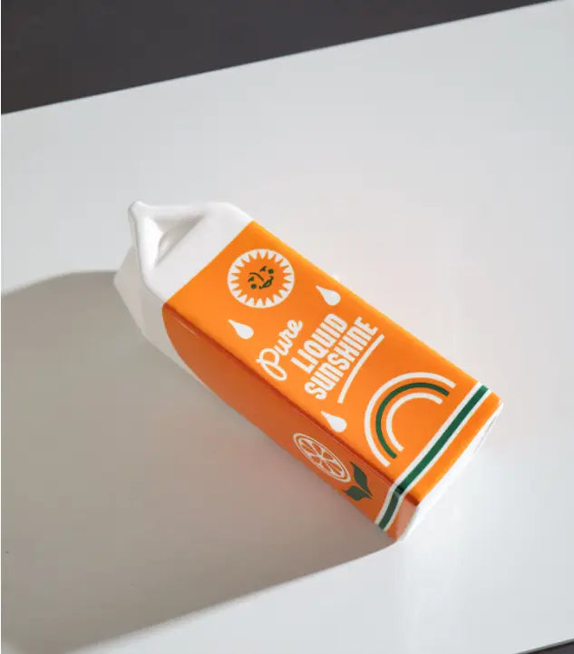 Vase en céramique en forme de brique de jus d’orange, en orange, avec un bouchon blanc et une base rayée verte, conçu pour ressembler à un jus d’orange « pure liquid sunshine ».
