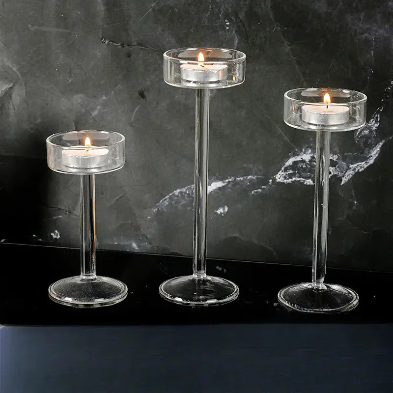 Tres portavelas de pilar de vidrio transparente, con tallos esbeltos y bases circulares, cada uno con una vela tipo té encendida, para una decoración moderna de mesa.