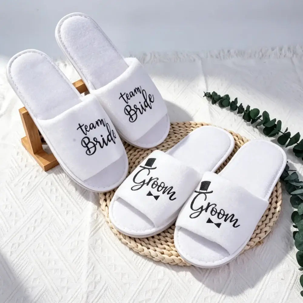 Weiße Slip-on-Slipper mit schwarzer Stickerei von Team-Bräute- und -Groom-Text sowie kleinen Fliege-Designs für das Schuhwerk der Hochzeitsgesellschaft.