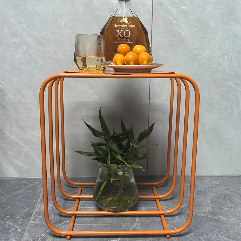Orange metal geometric side table with minimalist storage features a glass vase holding green plants on the lower shelf for decorative use.