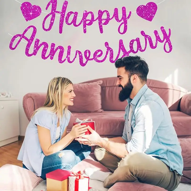 Red gift box with white ribbon shown alongside an anniversary banner party decoration glitter sign for festive celebrations.