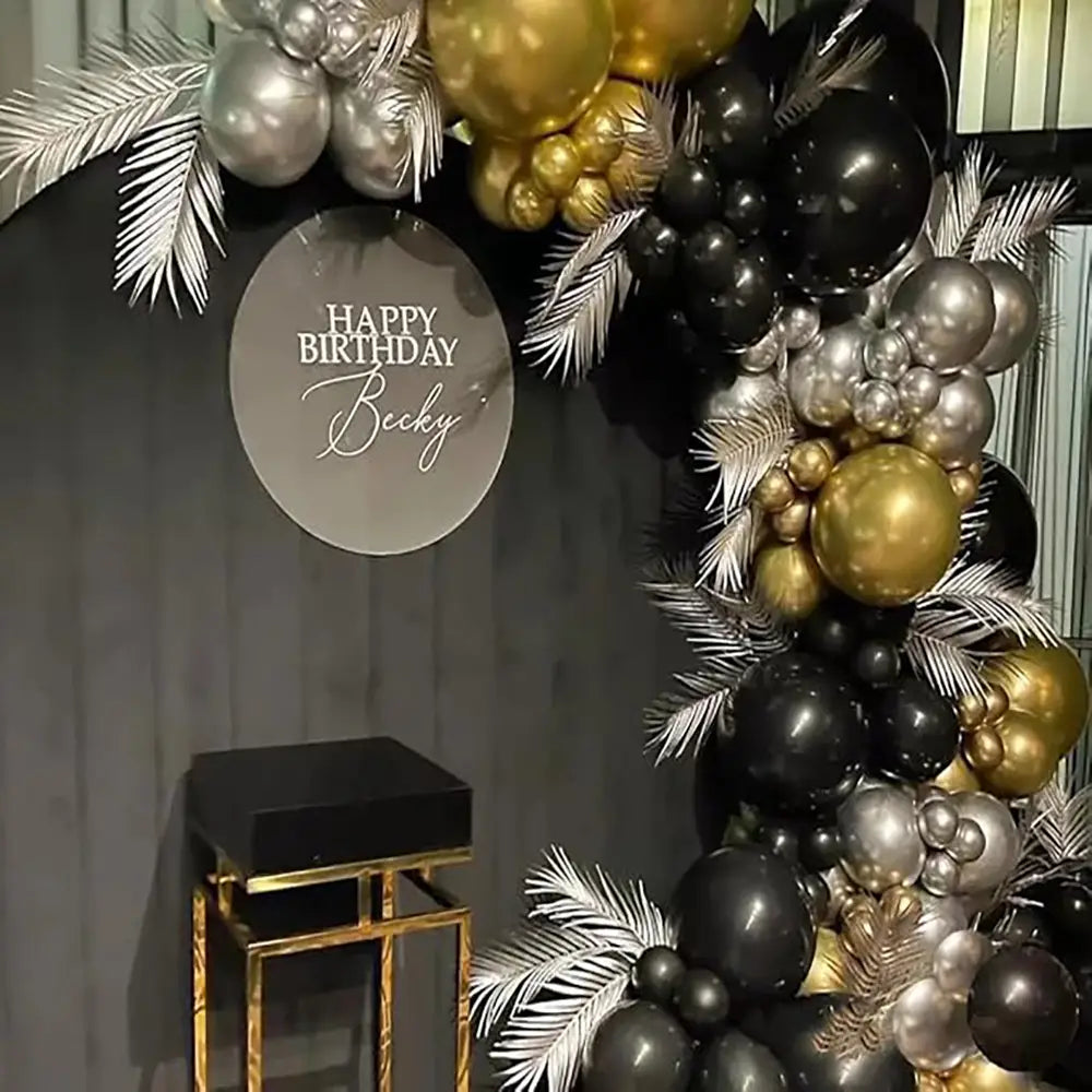 Schwarzes und goldenes Ballonbogen-Set mit metallischen schwarzen, goldenen und silbernen Ballons, verziert mit weißen Palmenwedeln, für Partys.