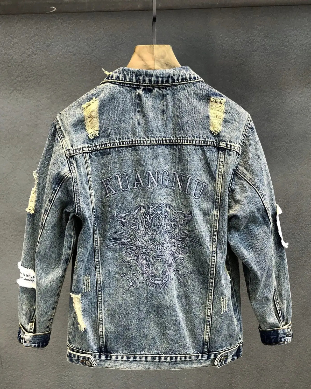 A distressed men’s embroidered tiger denim jacket in faded blue featuring ripped patches and a central tiger design above kuangniu lettering on the back.