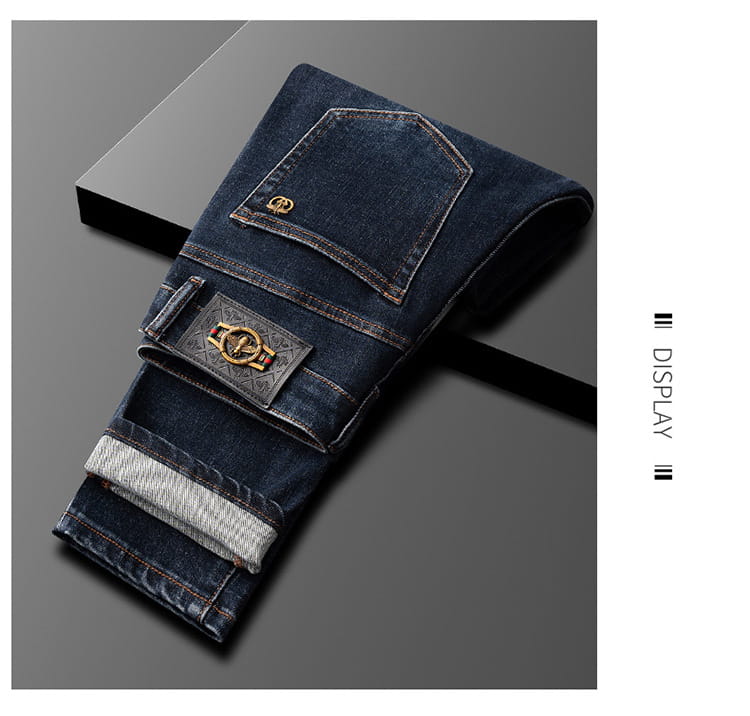 Slim fit dark blue denim jeans for men featuring a decorative gold toned metal emblem on the front pocket and contrasting light gray stitching.