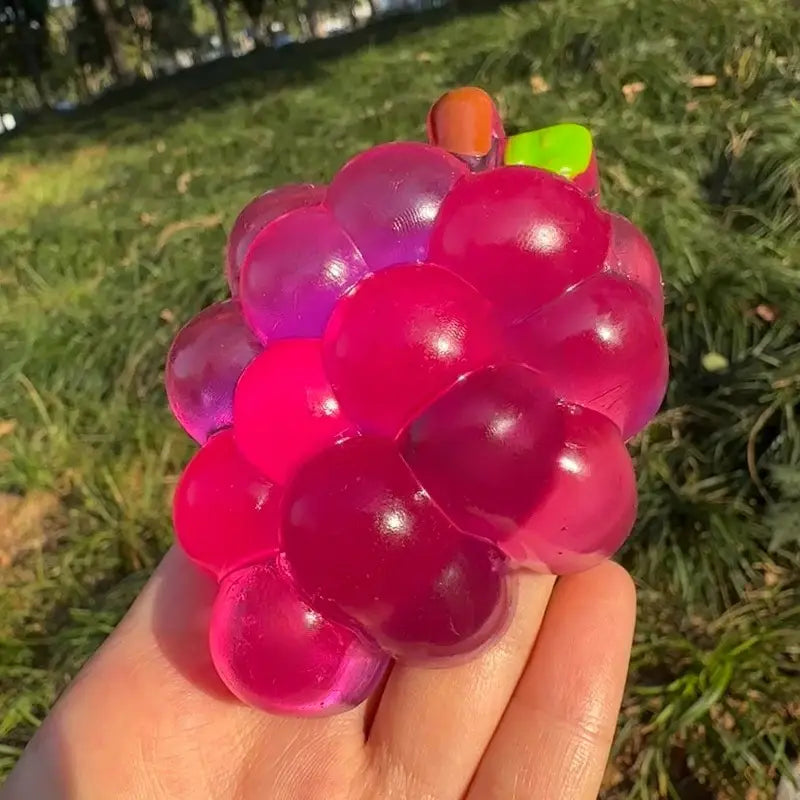 Ein transparenter, glänzender rosa- und violettfarbener Trauben-geformter Stressball, der in der Hand gehalten wird: Das Taba-Squishy-Grapefruit-Fidget-Spielzeug für sensorisches Taba-licious Fun Fruit-Spiel