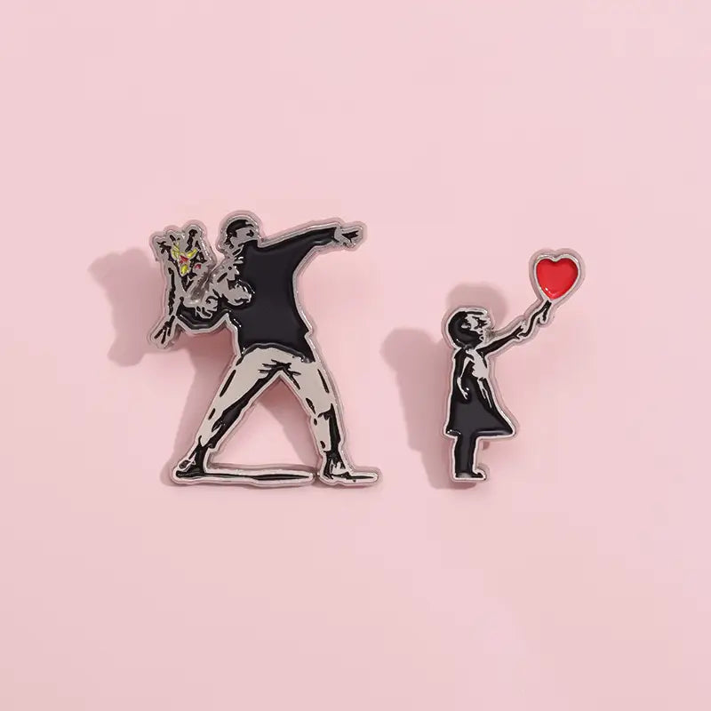 Deux épingles émaillées présentant des motifs d’art de rue emblématiques de Banksy : l’une en noir et blanc représentant un homme tenant des fleurs, et l’autre montrant un enfant avec un ballon en forme de cœur rouge.