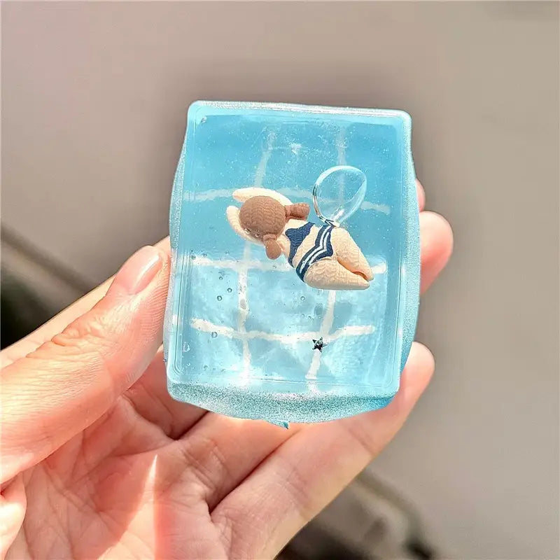 un cube sphérique translucide bleu à faible rebond, visqueux et lent, présentant une mini figurine de nageur portant un maillot à rayures bleu marine et blanc, flottant au sein d’une scène aquatique avec des lignes en quadrillage et de petites décorations en forme d’étoiles