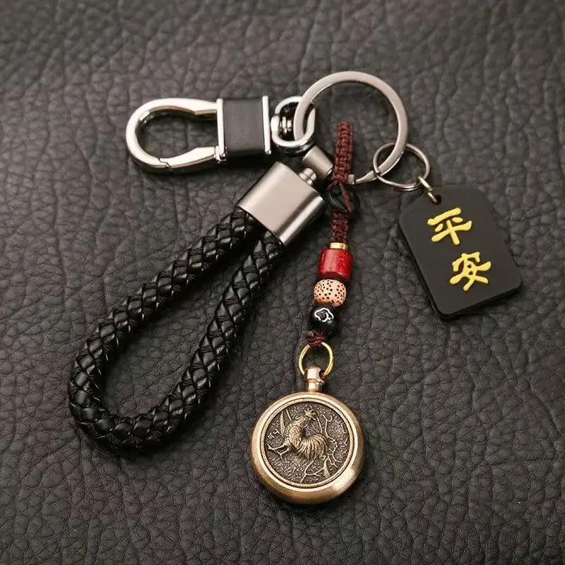 赤と黒のビーズでアクセントを施し、刻印された「鹿」のデザインをあしらったブロンズ色の円形ペンダントを備えた黒の編みレザー製キーチェーン。金色の漢字が記された小さな黒いタグ付き（中国の干支をモチーフにした編みレザー製ラッキーペンダント）。