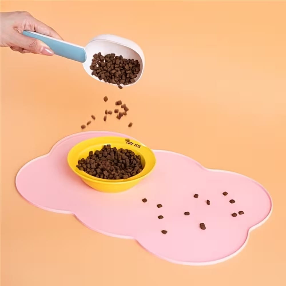 Alfombra de alimentación para mascotas de silicona con forma de nube color rosa para perros y gatos pequeños y medianos, con un cuenco amarillo y una pala con mango azul cerca