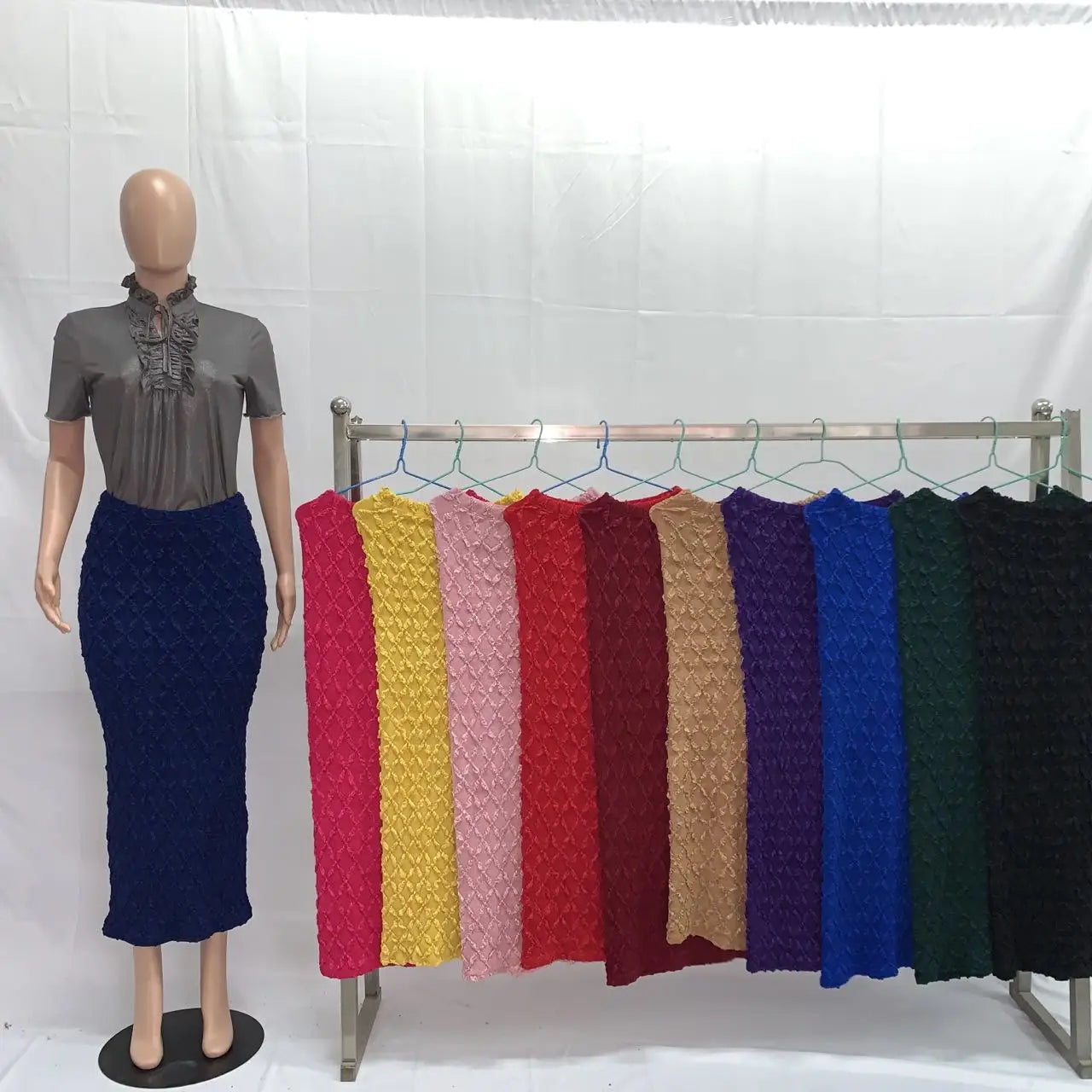 A mannequin wearing a gray ruffled blouse and a navy blue high waist knit midi skirt slim fit stands beside a rack displaying colorful pleated fabric skirts in various hues.