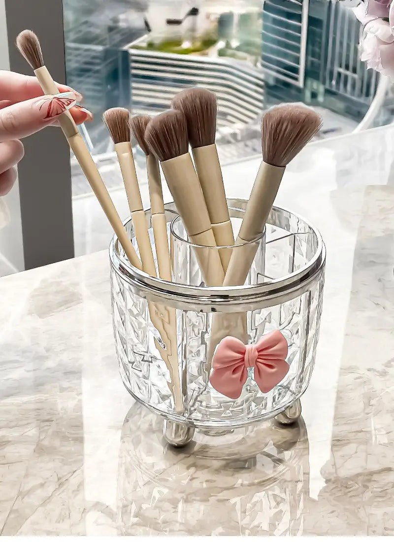 Porte-pinceaux en verre transparent avec accent de nœud rose et pieds argentés, dans une boîte de rangement tournante pour maquillage et organisation des cosmétiques pour le rangement des accessoires de maquillage.