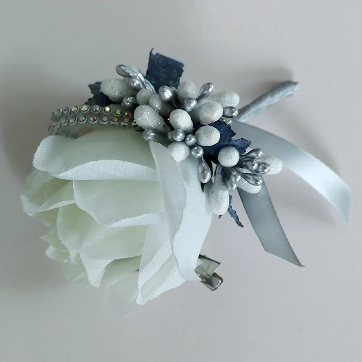 A delicate white artificial silk rose corsage with silver glittery beads, navy blue fabric accents, and a satin ribbon designed as a blue wedding boutonniere.