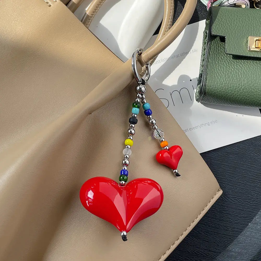 Porte-clés en forme de cœur rouge brillant, pendentif pour sac à perles, avec des perles colorées et des accents en argent, suspendus à une bandoulière de sac beige.