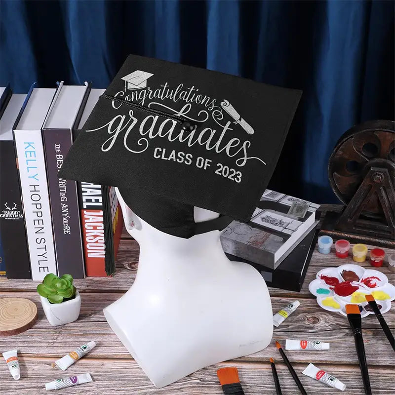 Black adult graduation cap with tassel featuring white embroidered congratulations graduates class of 2023 text and a small graduation hat icon.