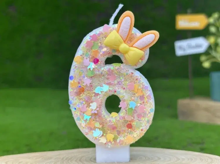 A translucent glittery number six birthday candle made of wax adorned with colorful confetti and topped with a yellow bow and bunny ears for birthday celebrations.