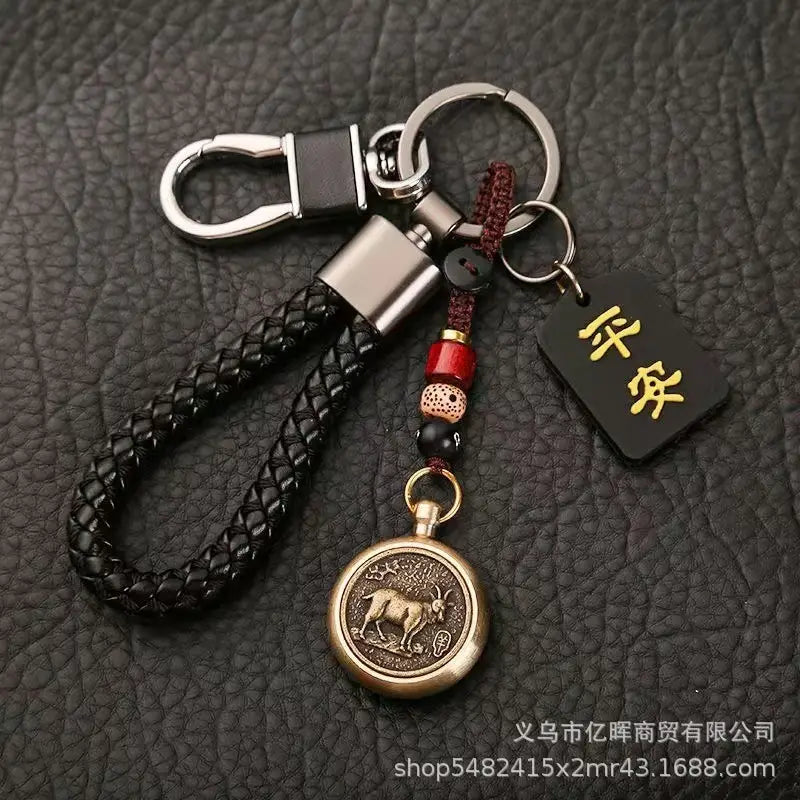赤と黒のビーズでアクセントを施し、ヤギの意匠を刻印したブロンズ色の円形ペンダントを備えた黒の編みレザー製キーチェーン。金色の漢字が記された小さな黒いタグ付き（中国の干支をモチーフにした編みレザー製ラッキーペンダント）。
