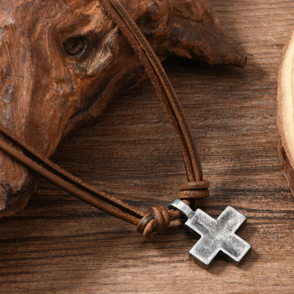 A rustic silver-toned alloy cross pendant with a textured surface hangs from a brown leather cord in a leather cross necklace.