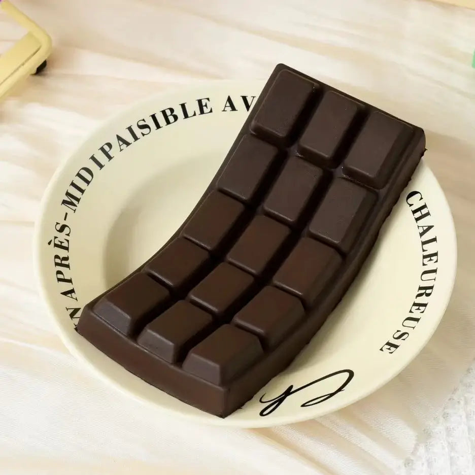 juguete exprimible con forma de barra de chocolate oscuro para aliviar el estrés, con acabado brillante y diseño segmentado, apoyado sobre un plato blanco