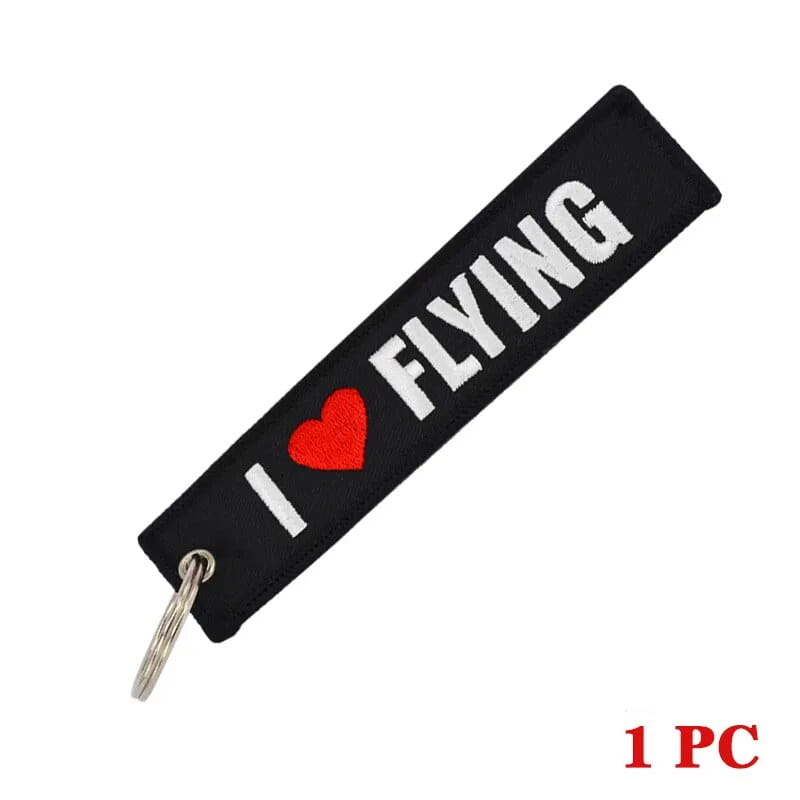 銀色の金属リング付き。白い「i heart flying」表記と赤いハートをあしらった黒い布地の「フライト前に取り外してください」刺繍キーチェーン。