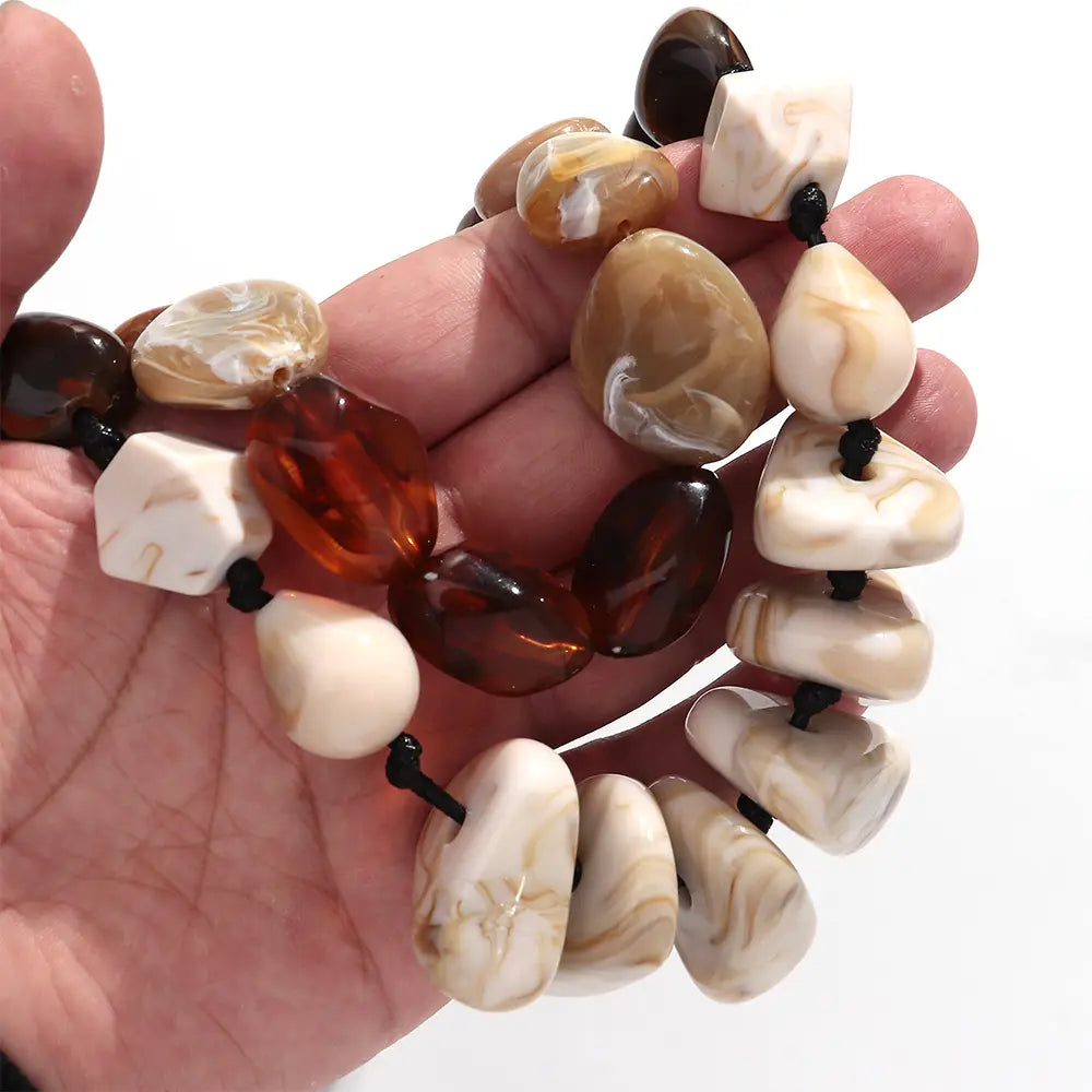 Une main tient un collier ras du cou en résine à motif géométrique, fait de perles marbrées, avec des perles en ambre et en agate polies de formes irrégulières, dans des tons chauds brun, blanc crémeux et orange translucide.