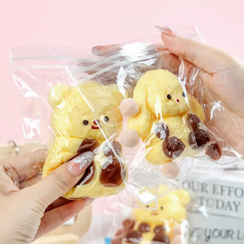 Gelbe Plüschbären-förmige Quetschspielzeuge mit braunen schokoladenartigen Akzenten und pinken Pompons auf den Wangen, verpackt in einem durchsichtigen Plastiksäckchen zur sensorischen Ablenkung und zum Stressabbau.