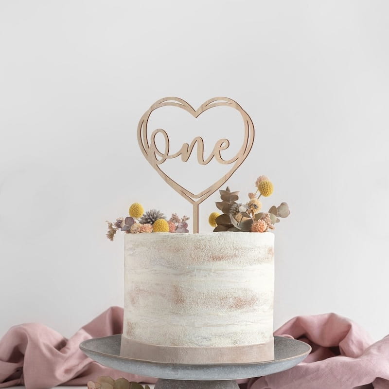 Un topper de gâteau en bois en forme de cœur doré, avec le mot « one » découpé en écriture cursive, pour une décoration florale d’anniversaire