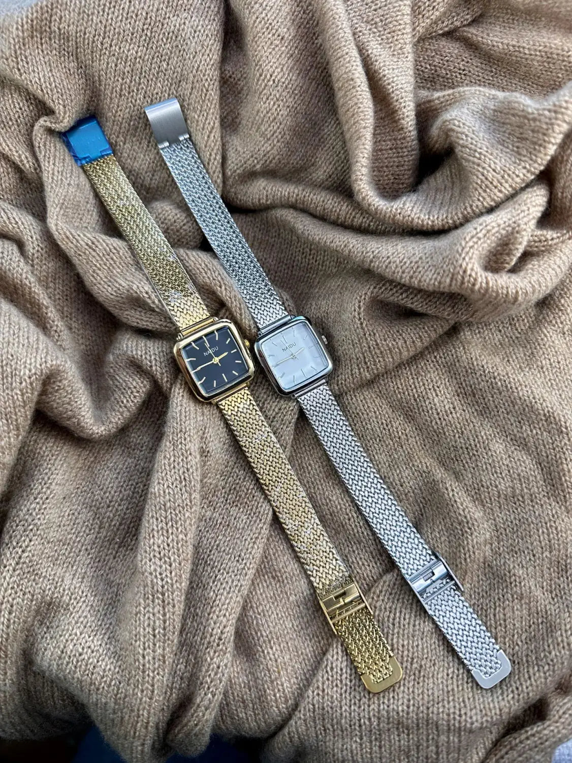 Two elegant minimalist square quartz watches for women with mesh bands in gold tone and silver tone resting on soft beige knitted fabric.