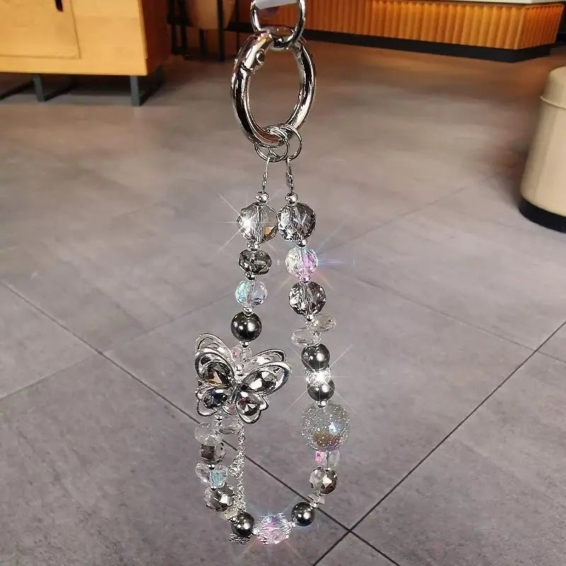 Une chaîne pour téléphone mobile à breloque papillon argentée, ornée d’une cascade de perles transparentes et irisées, destinée à servir de bracelet de téléphone mobile décoratif.