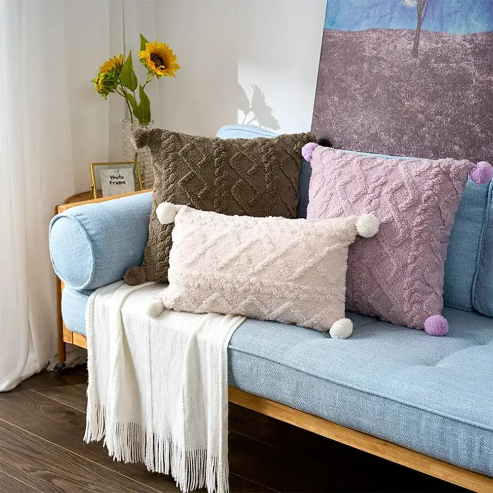 Three plush geometric throw pillow covers 45x45cm in muted brown soft lavender and creamy off-white with textured fabric and fluffy pom-poms at the corners.