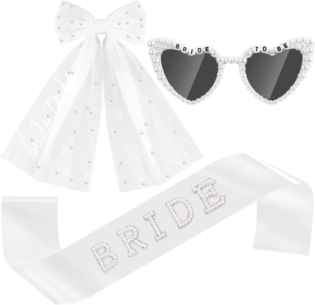 Conjunto de accesorios nupciales en blanco que presenta un lazo con cuentas, gafas de sol con forma de corazón con la inscripción «bride to be», y una faja adornada con pedrería, diseñada para celebraciones nupciales.
