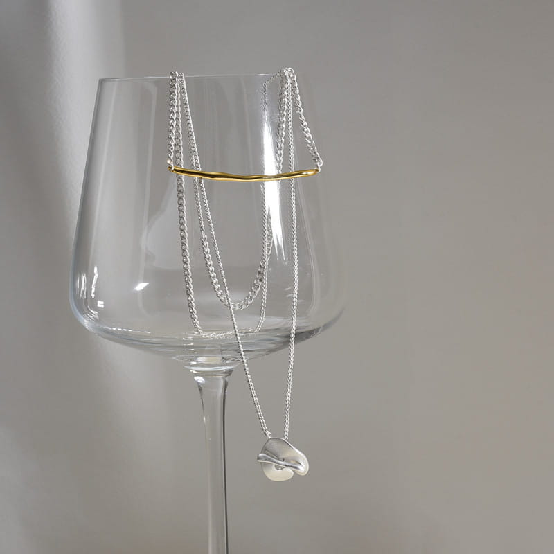 Collier délicat à chaîne en argent pour femmes, présentant un pendentif abstrait en forme de cœur minimaliste suspendu avec élégance au bord d’un verre à vin transparent.