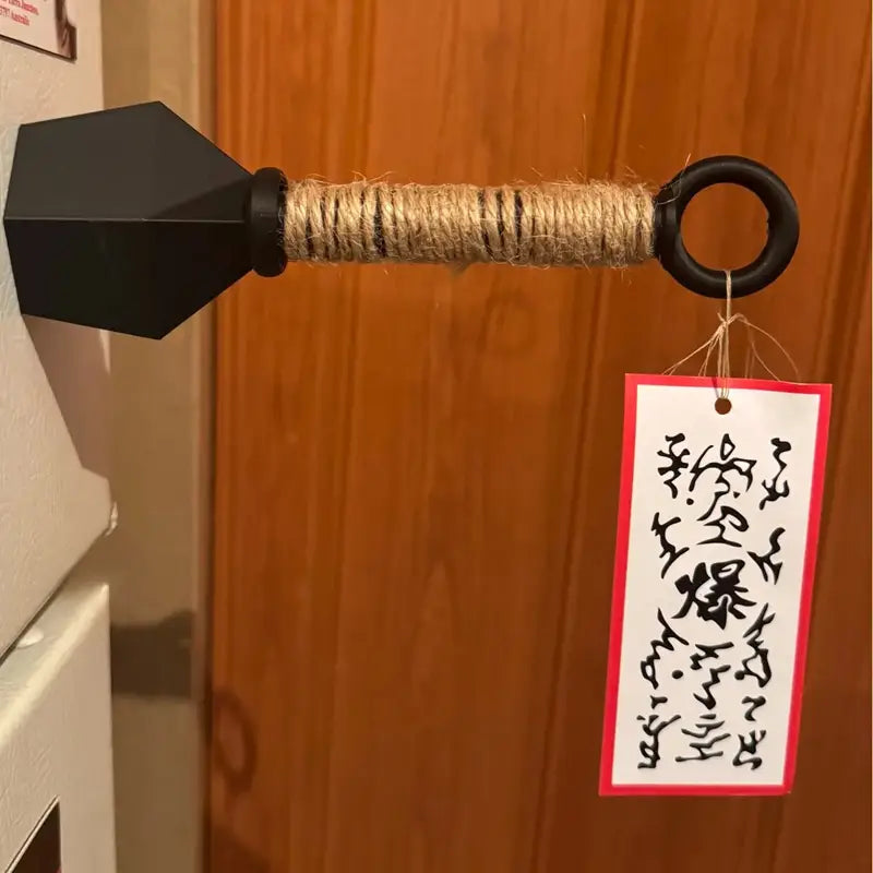 schwarzer Türgriff in Pfeilform mit strukturierter, natürlicher, mit Bindfaden umwickelter Schaftpartie und anhängendem Schild, dargestellt als Teil eines Ninja-Kunai-Kühlschrankmagneten aus PVC, Shuriken-Spielzeug.