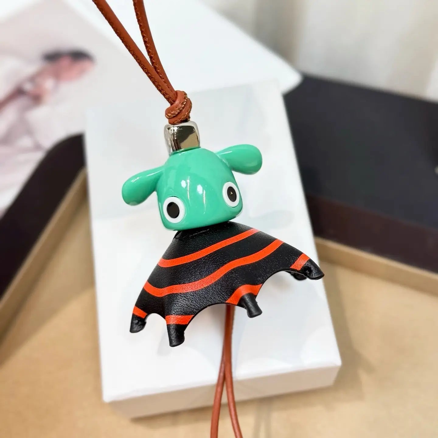 Llavero colgante para bolso con encanto de animales de cuero, tierno, de diseñador, con un murciélago de cerámica turquesa, alas de cuero a rayas negro y naranja, sujeto a un cordón de cuero marrón.