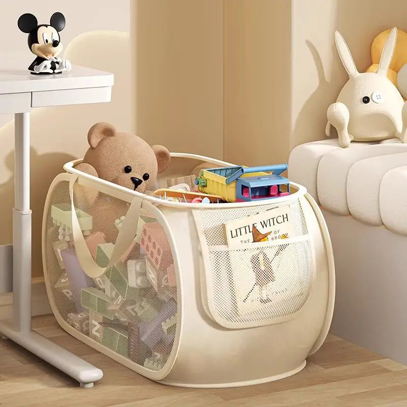 Cream-colored mesh toy storage basket with large capacity and front pocket holding a little witch book for effective toy organization.