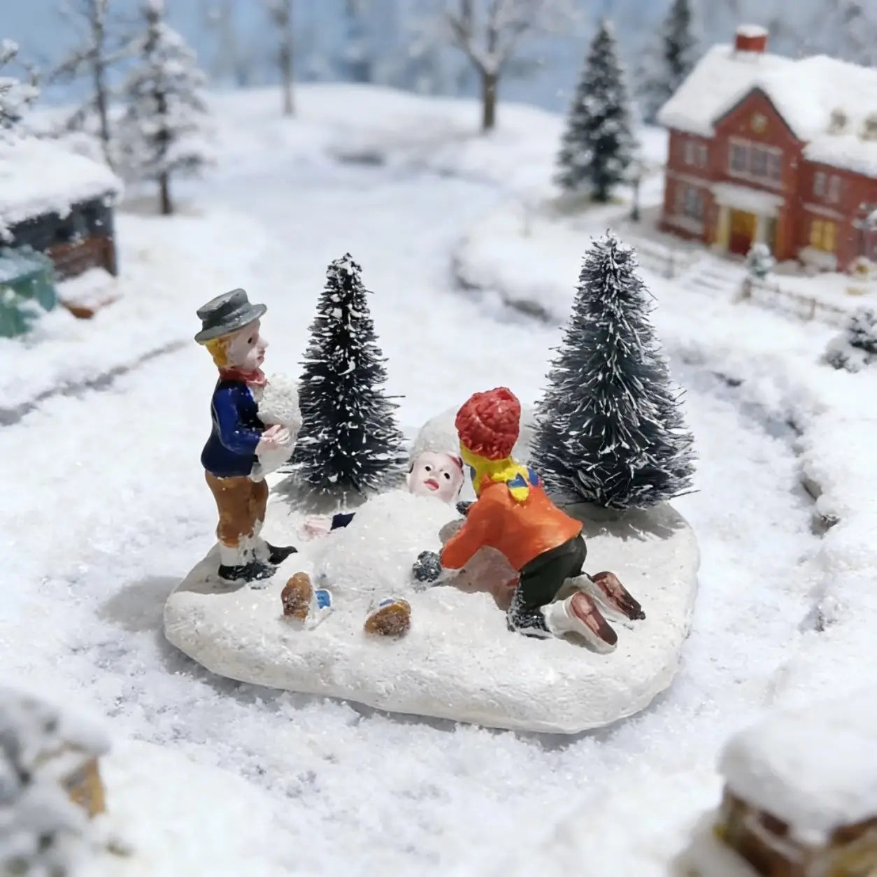 Kinder spielen mit Schnee – Weihnachtliches Dorf-Figürchen: mit einer Miniatur-Schneeszene, zwei Kindern und einem Schneemann auf einer verschneiten Basis, mit kleinen Bäumen und Häusern im Hintergrund.