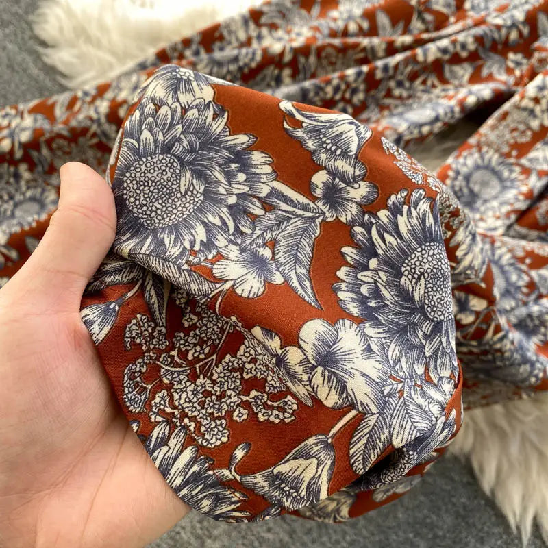 Una mano sostiene una tela color naranja óxido suave con un intrincado estampado floral negro y blanco, con girasoles detallados, con escote tipo bardot en un enterizo de pierna ancha.