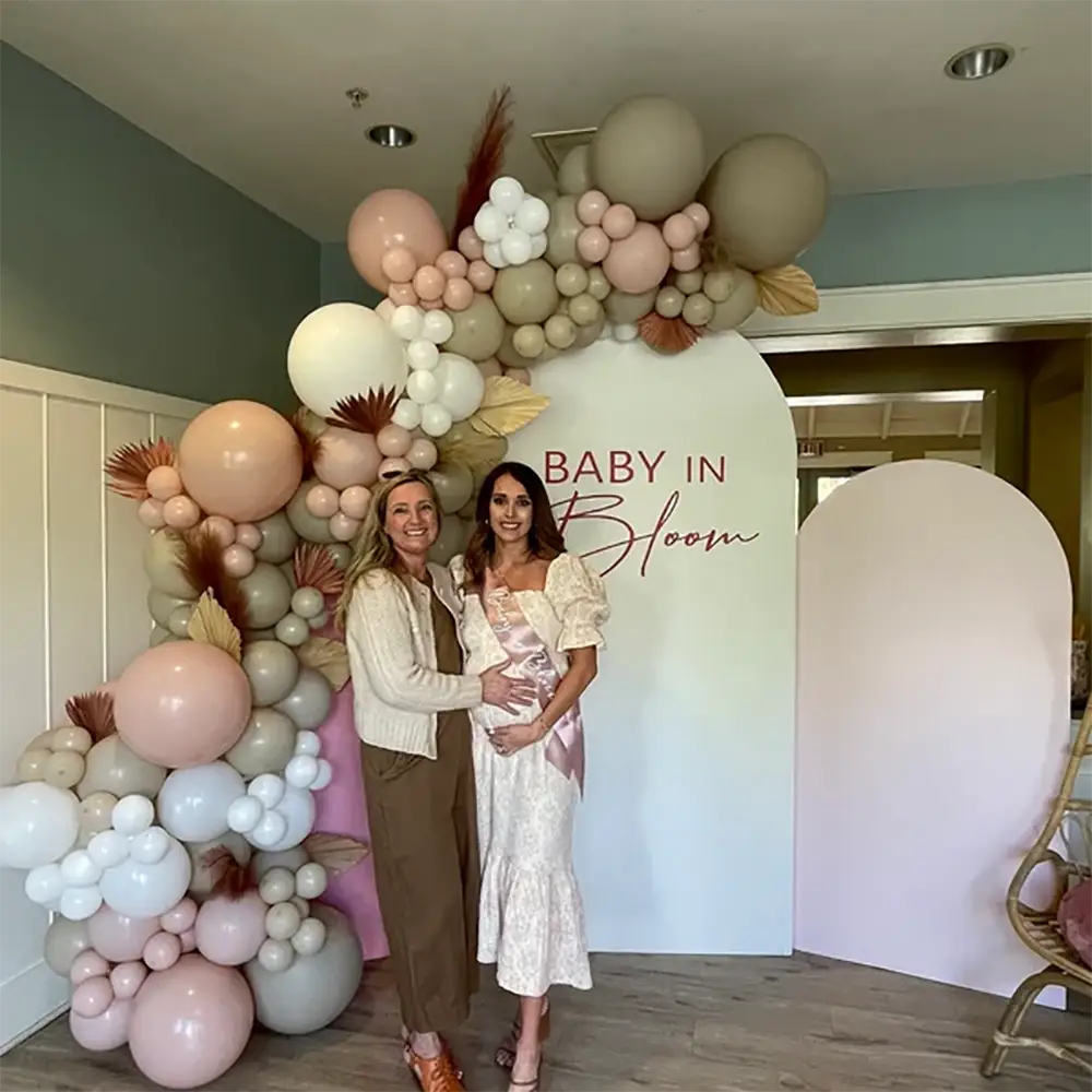 Une écharpe de baby shower en satin blush rose pâle avec un motif « mum » présenté aux côtés d’une arche de ballons rose tendre et crème, agrémentée d’accents de pampas séchées.