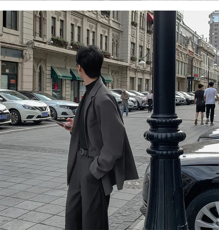 Un hombre que lleva una chaqueta de traje de doble botonadura entallada de poliéster gris, con cuello vuelto negro y cinturón a juego, de pie en una acera de la ciudad.