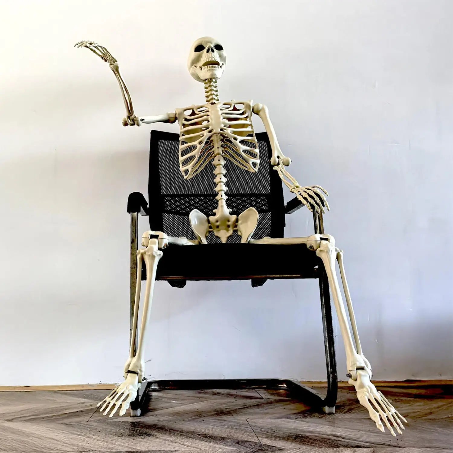 una decoración posable de esqueleto de Halloween de plástico color beige, sentada en una silla de oficina de malla negra, para exhibiciones de casas encantadas